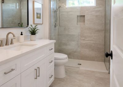 Modern bathroom with white vanity, glass shower, toilet, beige tile floor, and a small potted plant on the counter.