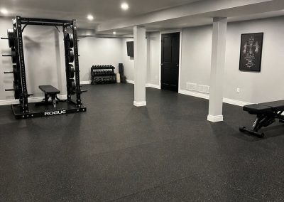 Home gym with weight rack, bench, dumbbells, wall poster, and rubber flooring in a spacious room.