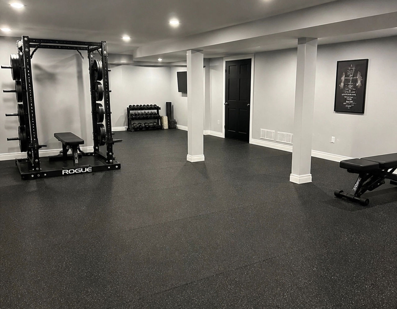 Home gym with weight rack, bench, dumbbells, and light gray walls; rubber flooring throughout the space.