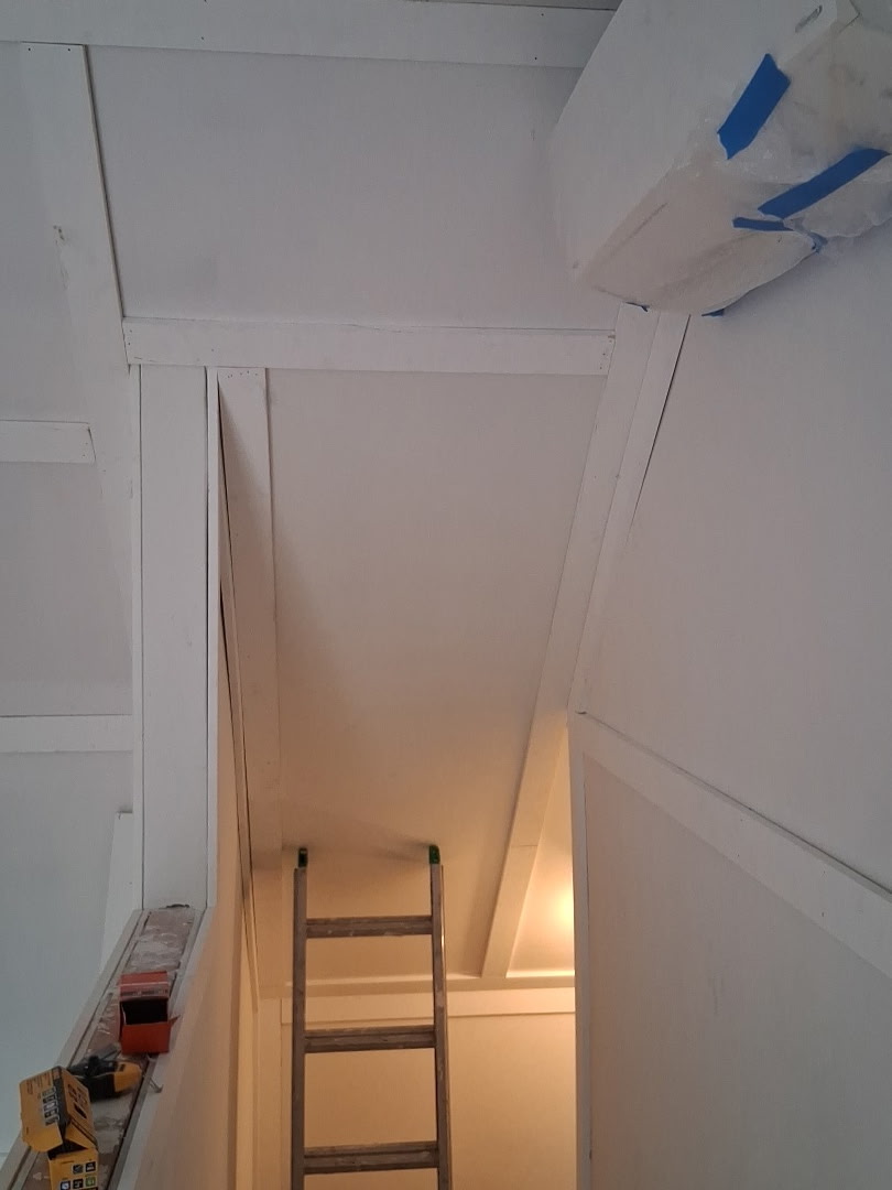 A wooden ladder leans against a corner wall in a room under construction with white walls and beams.