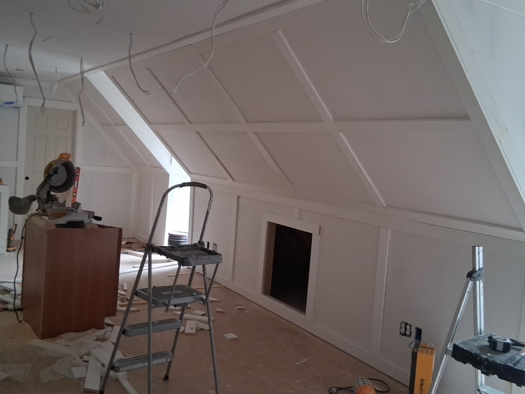 Unfinished attic room with white paneled walls, power tools, ladder, and hanging electrical wires.