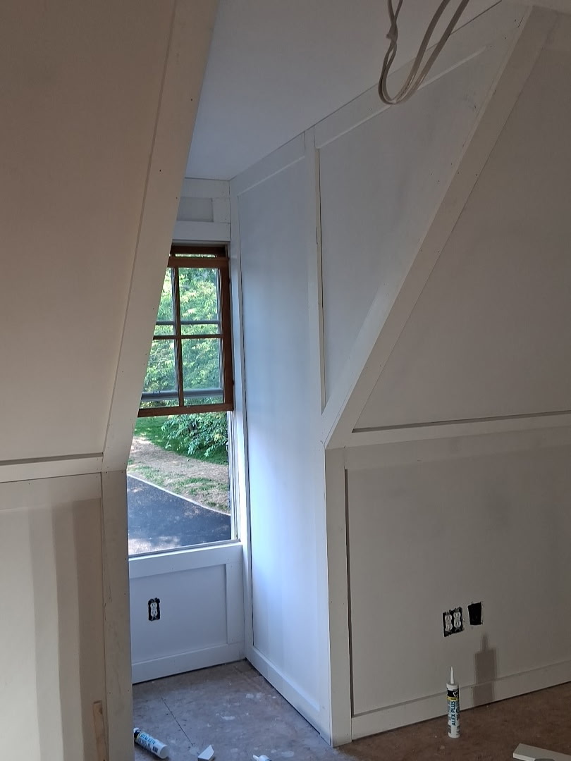A white-painted room with slanted walls, a window, and electrical outlets; renovation supplies are on the floor.