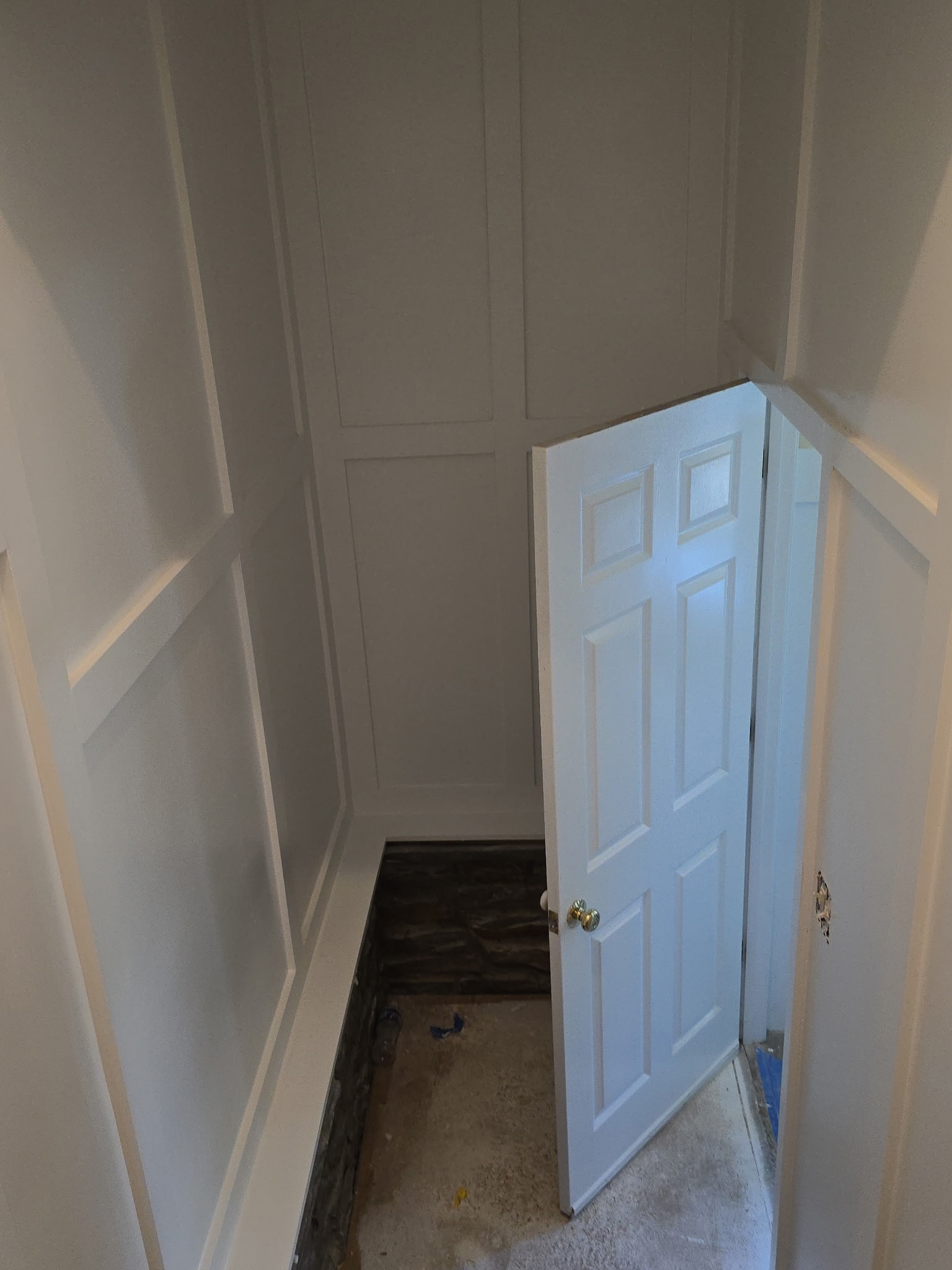 White paneled room with a partially open door and an unfinished floor.