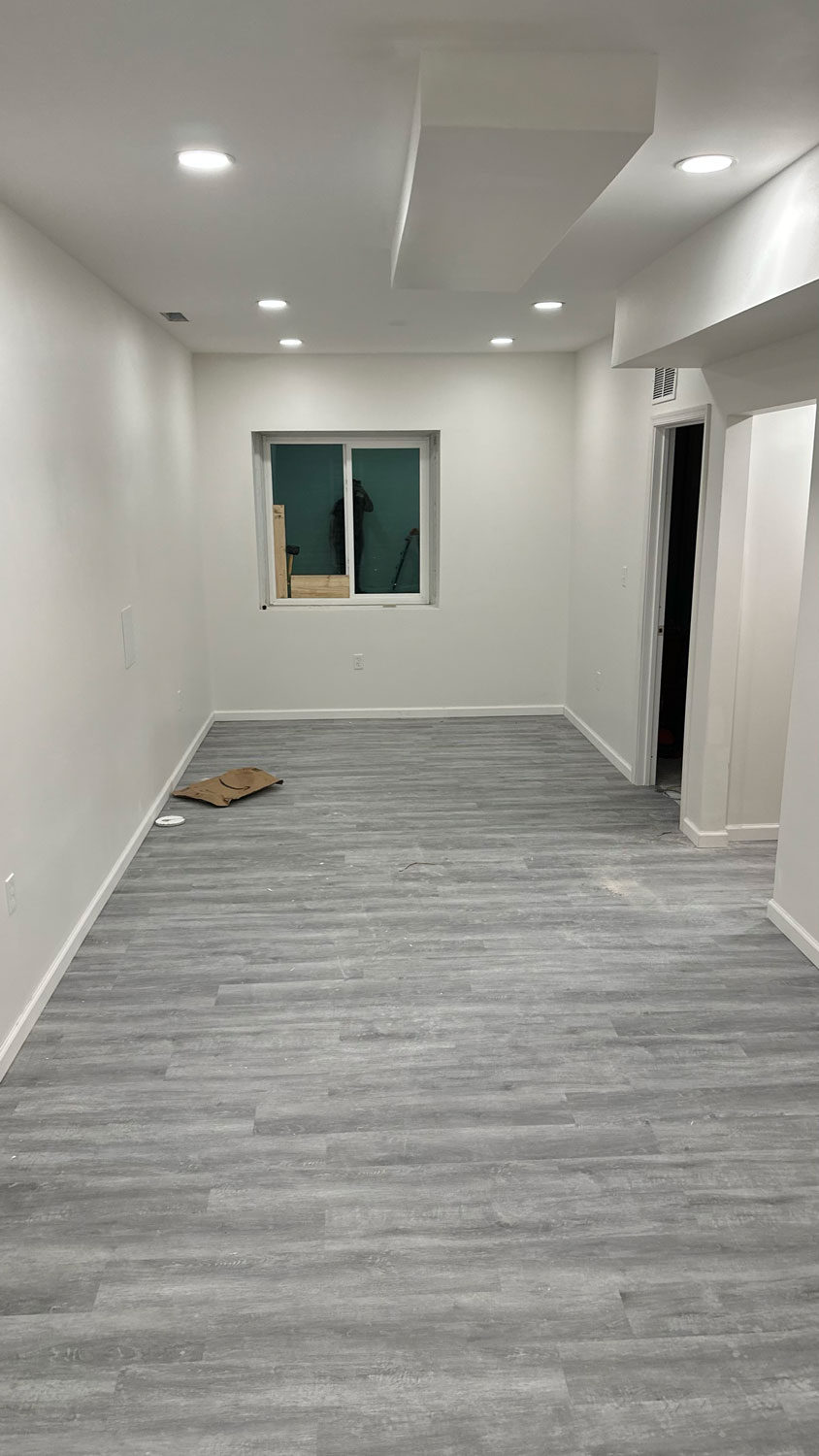 A long, empty room with gray wood floors, white walls, and a window at the far end.