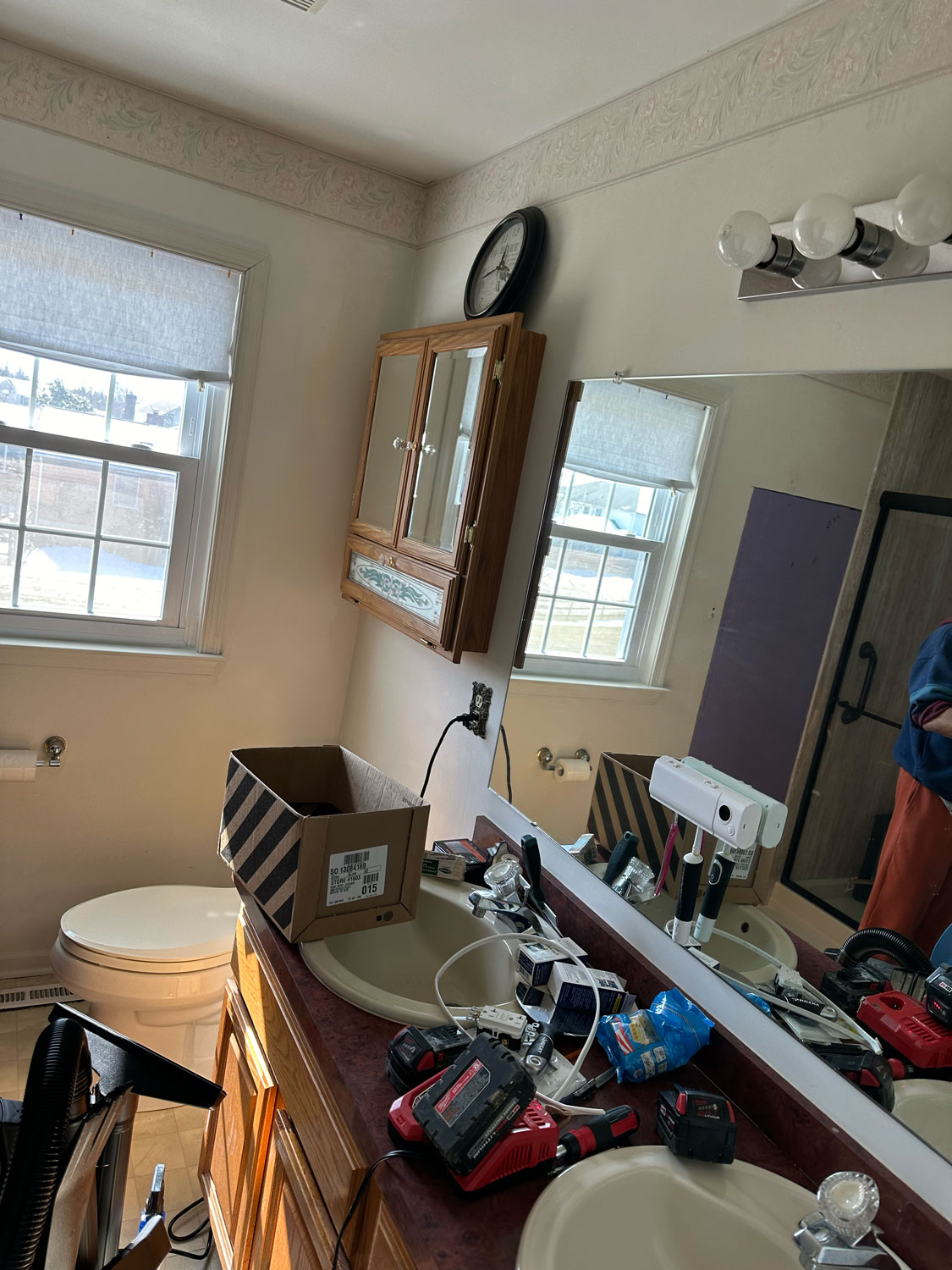 Cluttered bathroom with tools and a box on the counter, well-lit by natural light from a window.