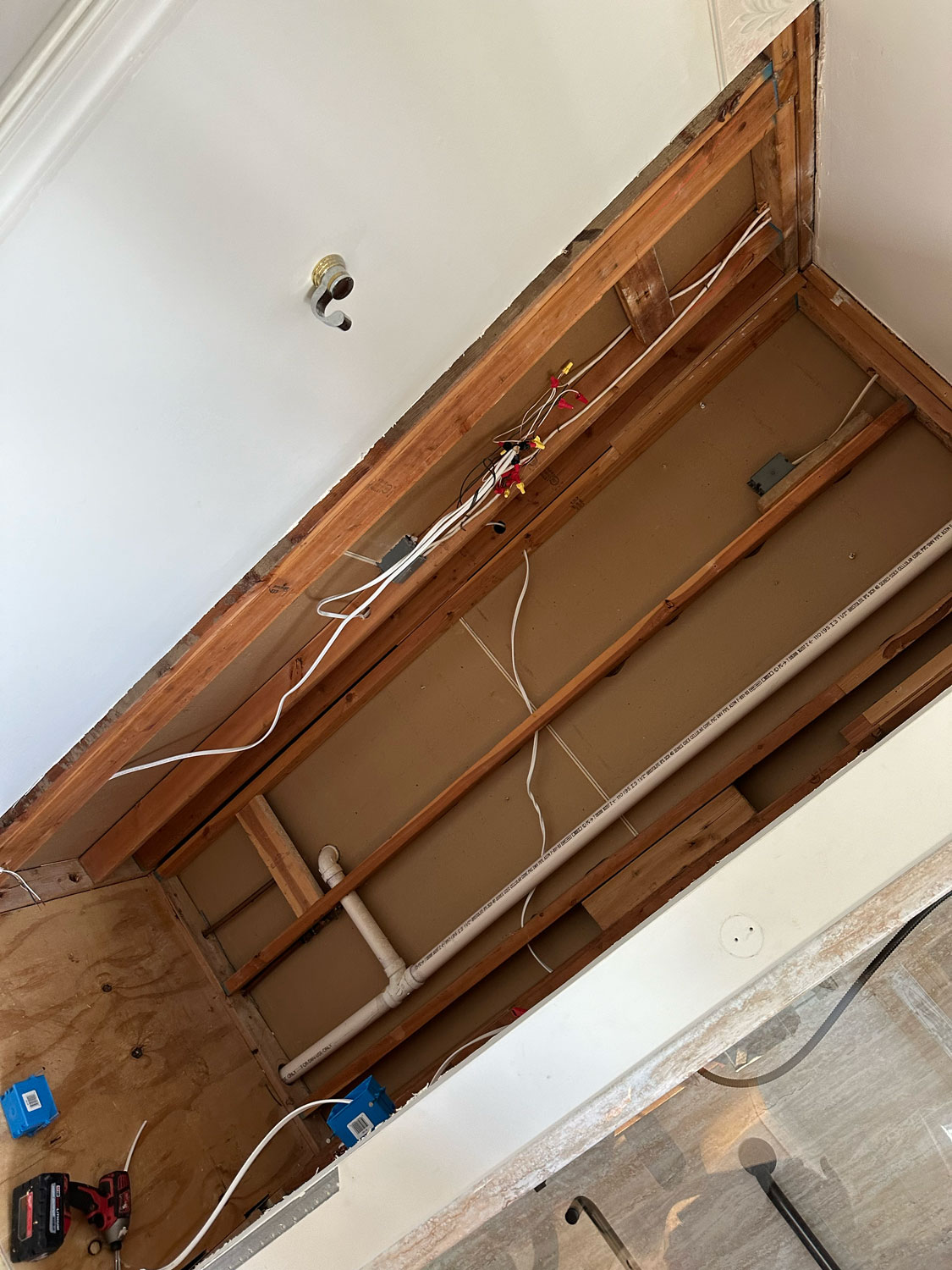 Ceiling partially removed, exposing wooden beams, electrical wiring, and plumbing pipes in a room under renovation.