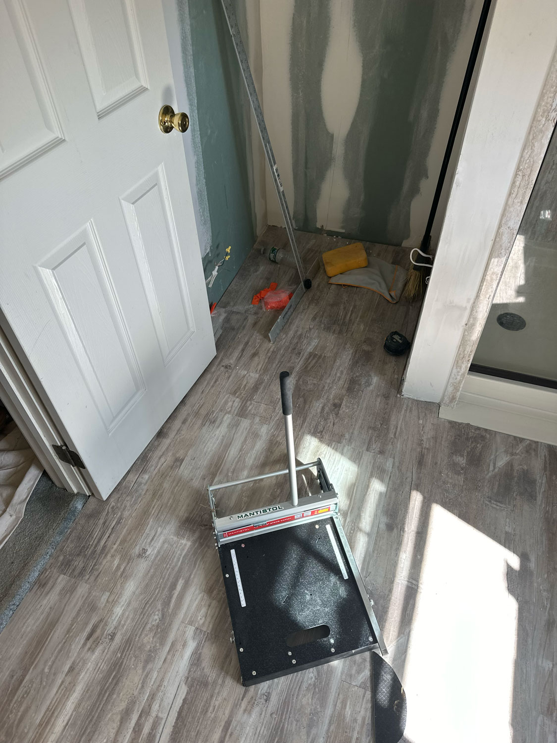 A flooring cutter, tools, and supplies are scattered on the floor of a room under renovation.
