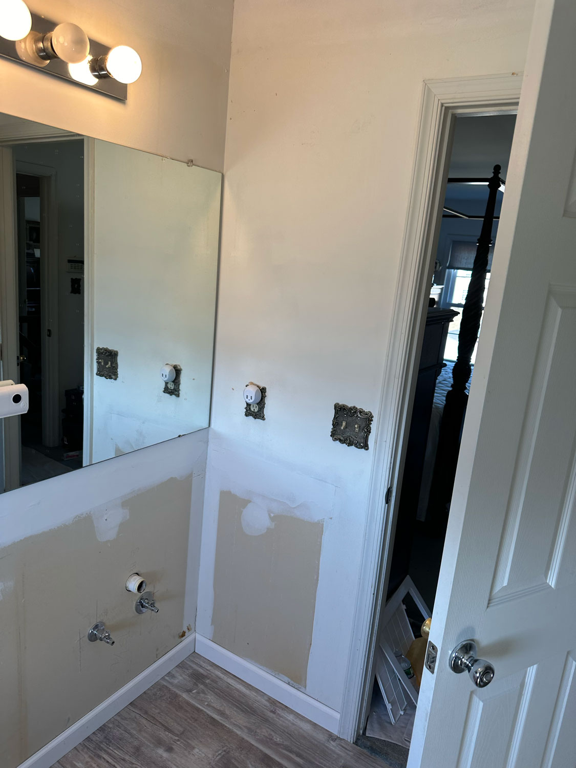 Bathroom under renovation with wall patches, exposed plumbing, and missing outlets below a large mirror.