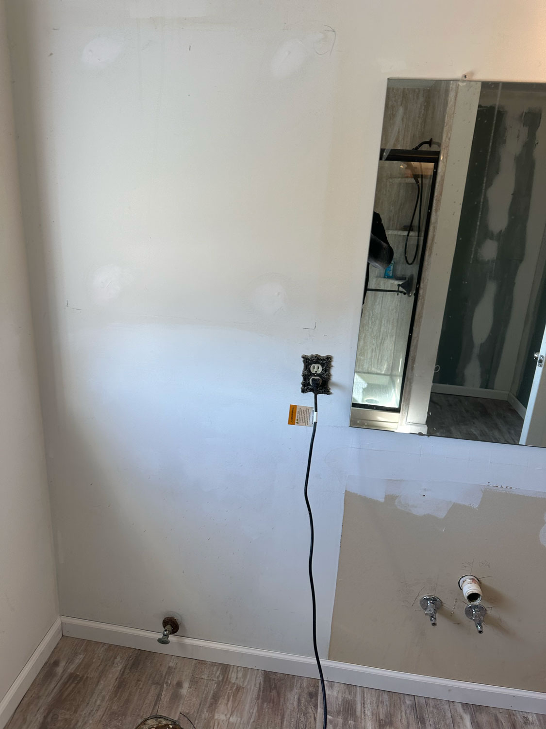 Partially renovated bathroom wall with exposed pipes, outlet, and unfinished drywall near a mirror and shower.