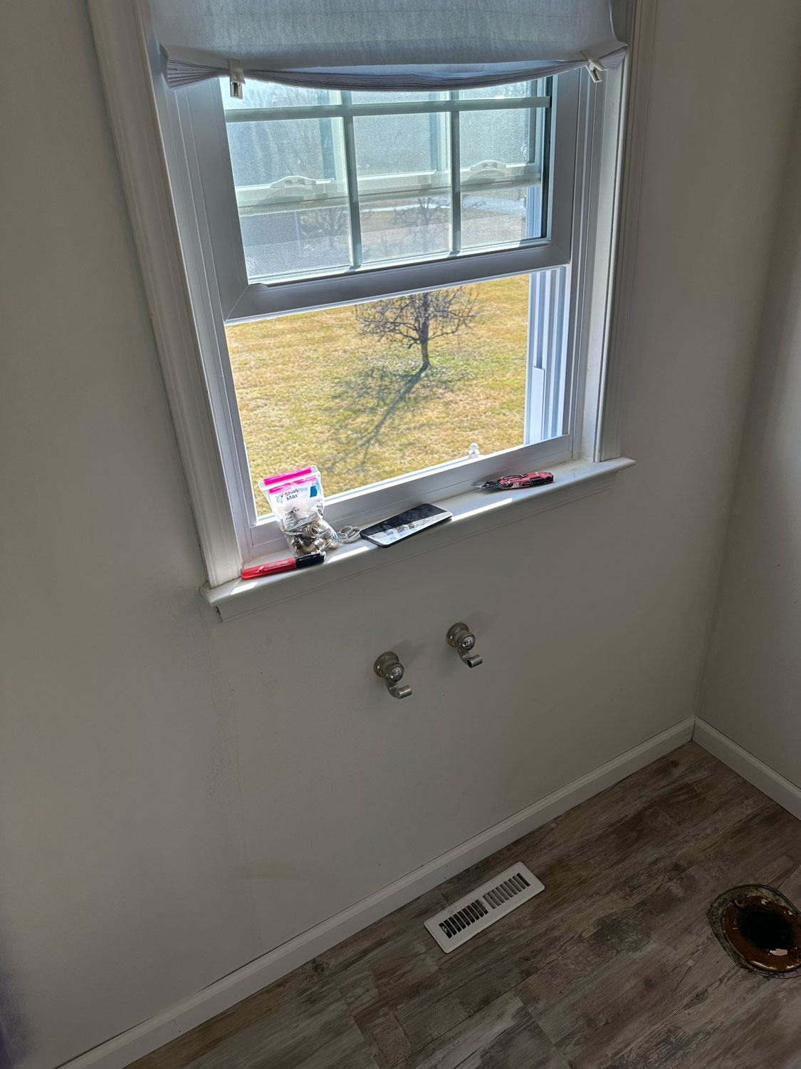 A small window above two exposed water pipes in an empty room with a vent and wood-patterned floor.