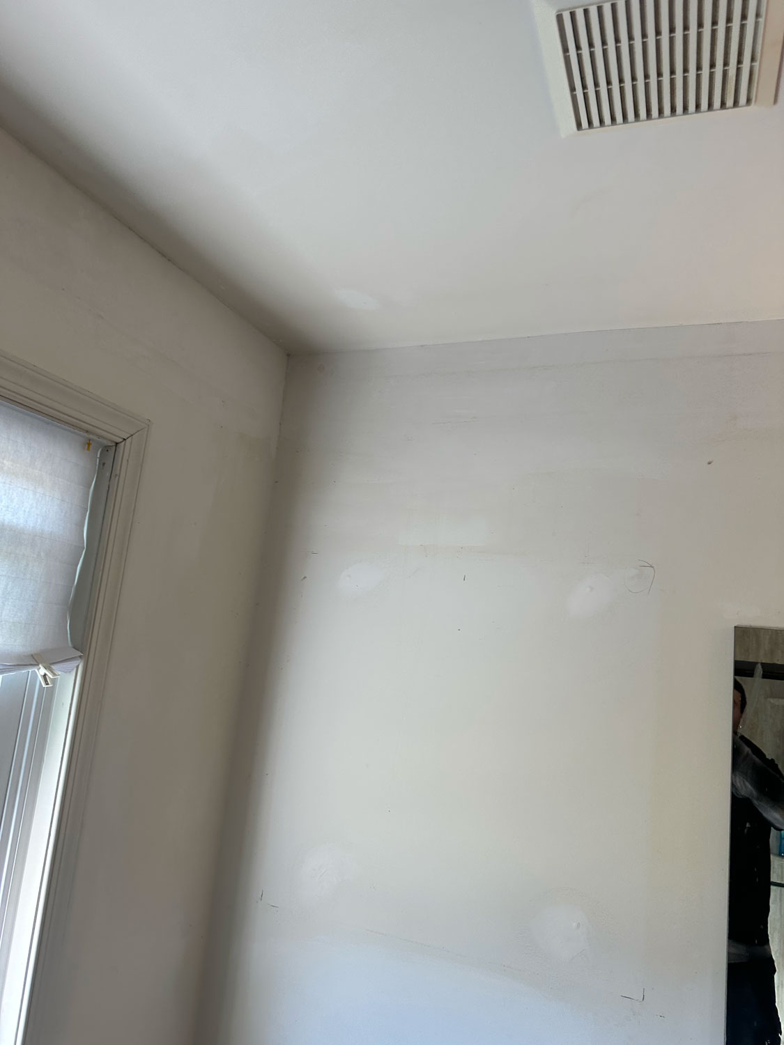 White wall and ceiling corner with patched spots, vent on ceiling, window and mirror partially visible.