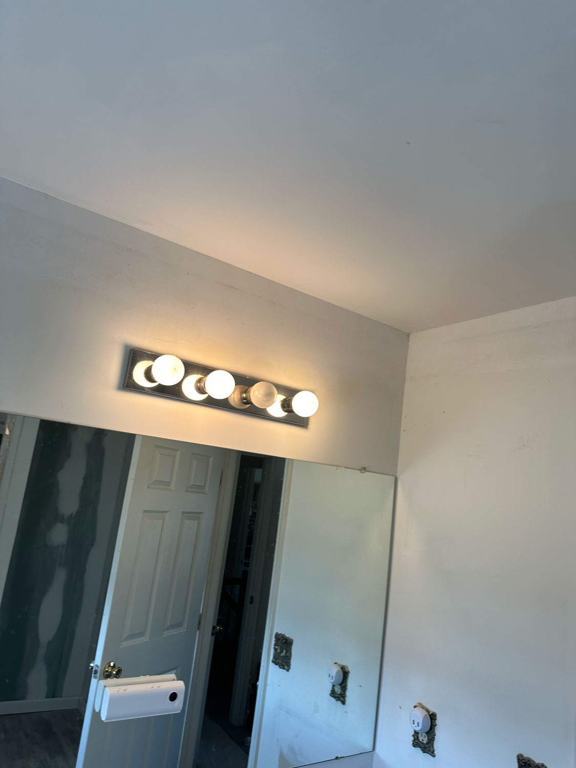 Bathroom wall with a mirror, a row of four lit vanity lights, and unfinished outlets without covers.