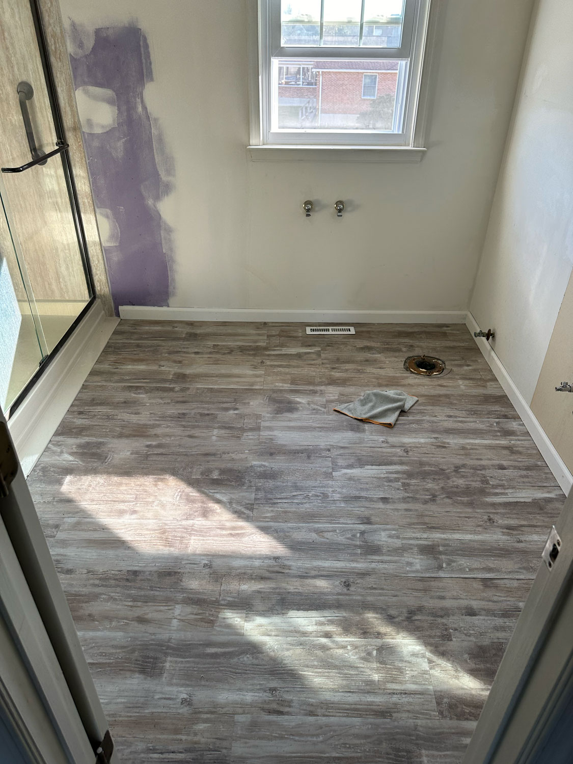 Bathroom under renovation with new wood-look flooring, exposed plumbing, and a patch on the floor.