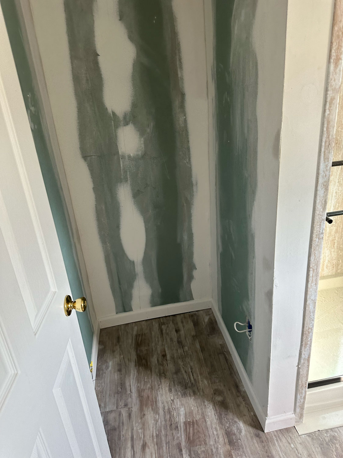 A small unfinished room with drywall seams exposed and wood-look flooring, viewed from an open door.