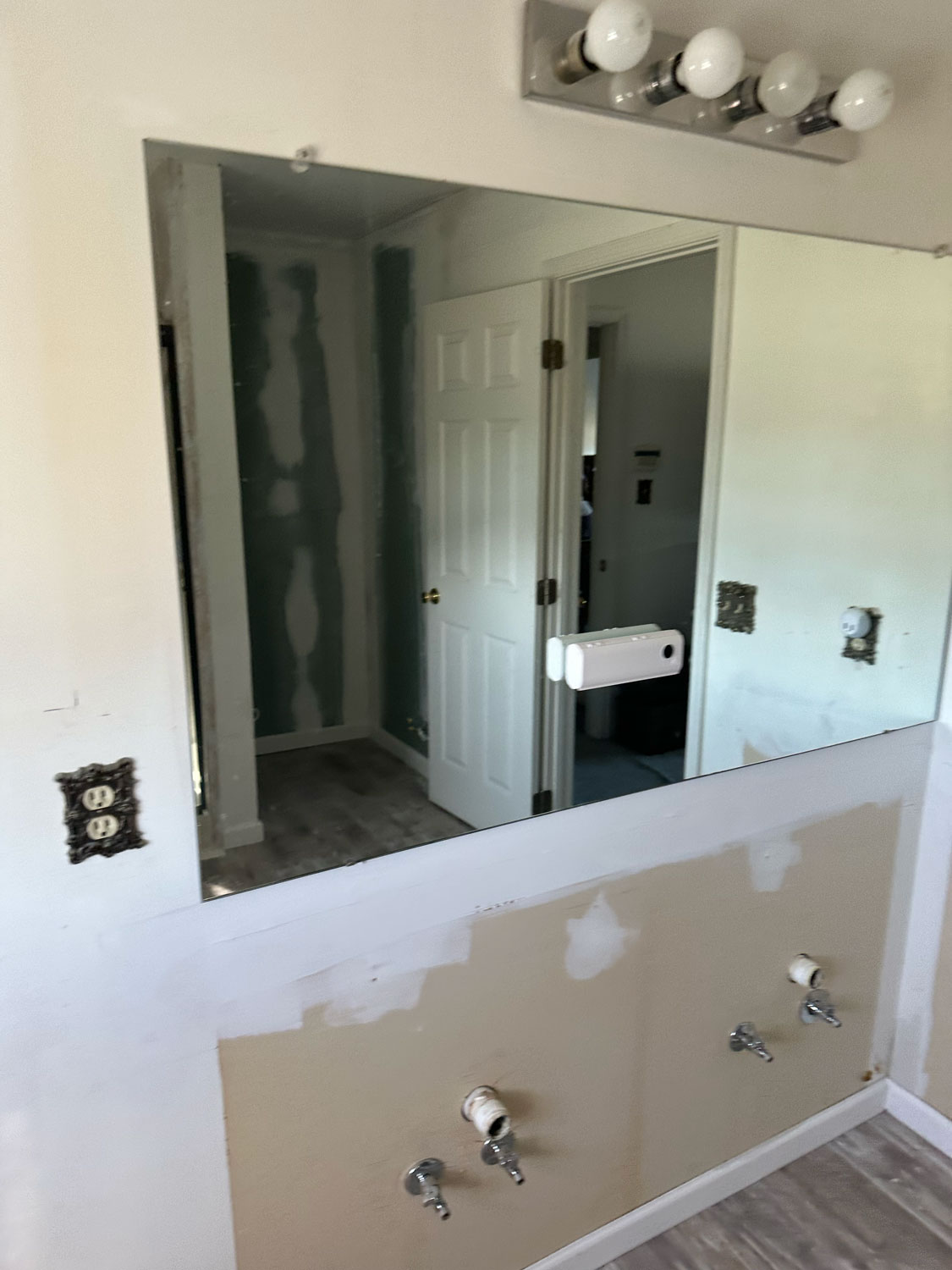 A bathroom under renovation with exposed plumbing, missing sinks, and an unfinished wall reflected in the mirror.