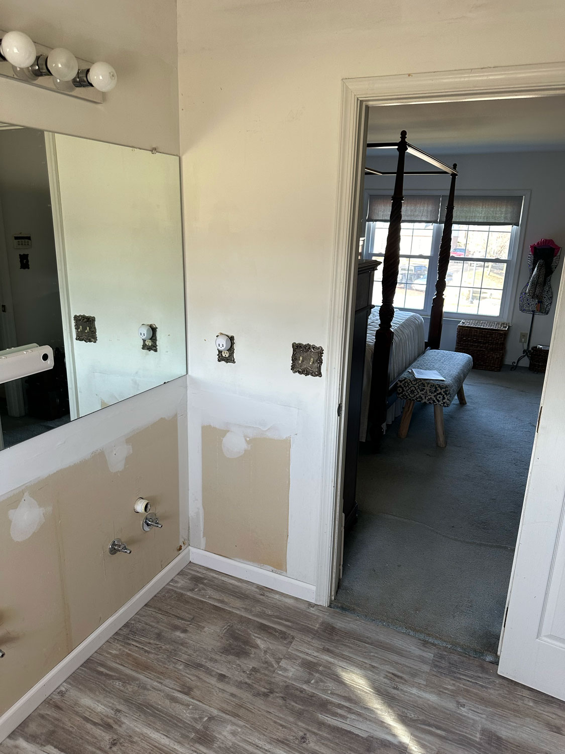 Bathroom under renovation with missing sink, exposed pipes, and a view into a bedroom with a four-poster bed.