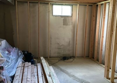 Unfinished basement room with exposed wooden studs, window, lumber, and construction materials on the floor.