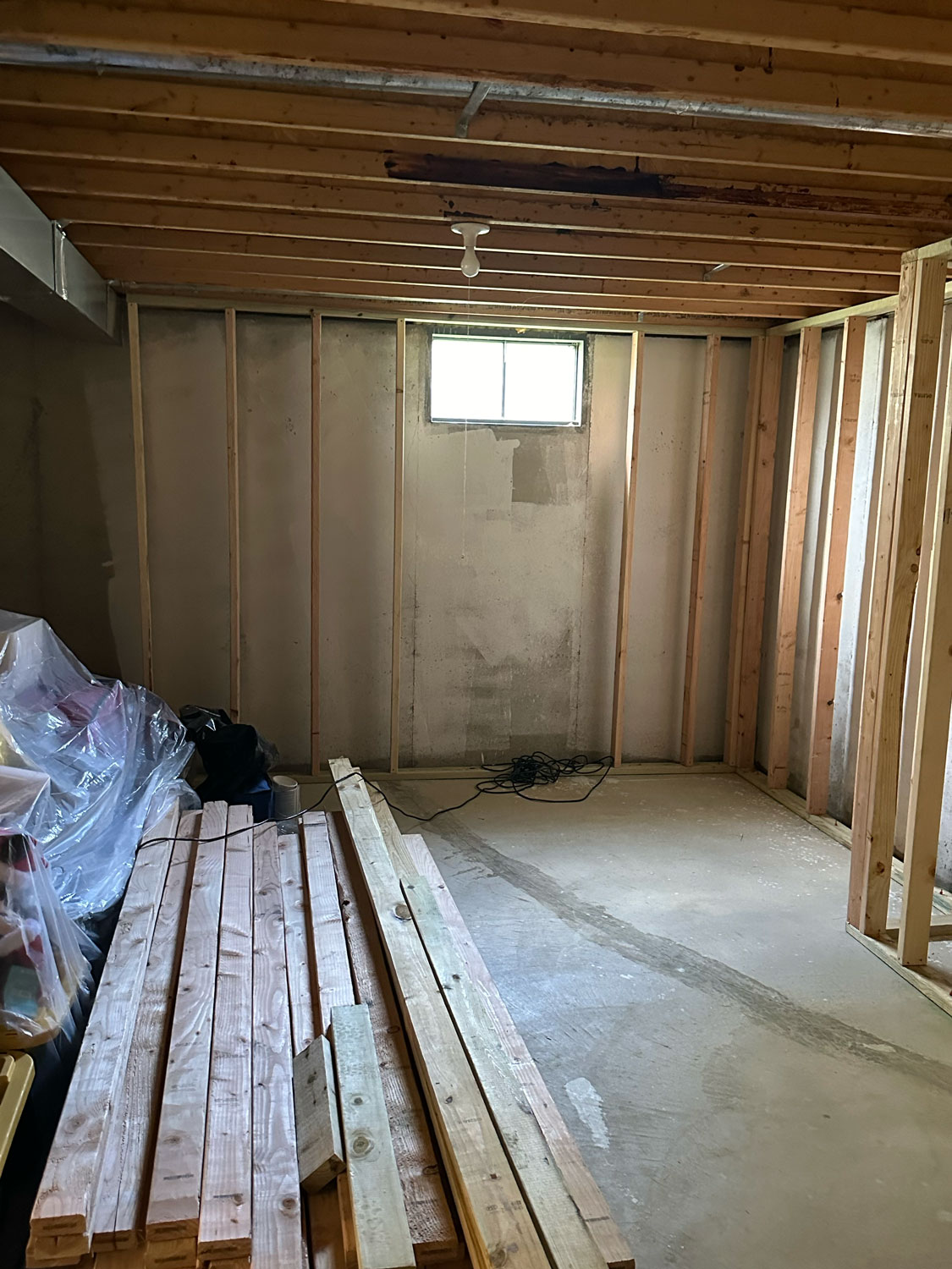 Unfinished basement room with exposed wooden studs, window, lumber, and construction materials on the floor.