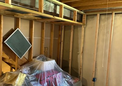 Unfinished basement with exposed wooden beams, stored items covered in plastic, and a hanging lightbulb.