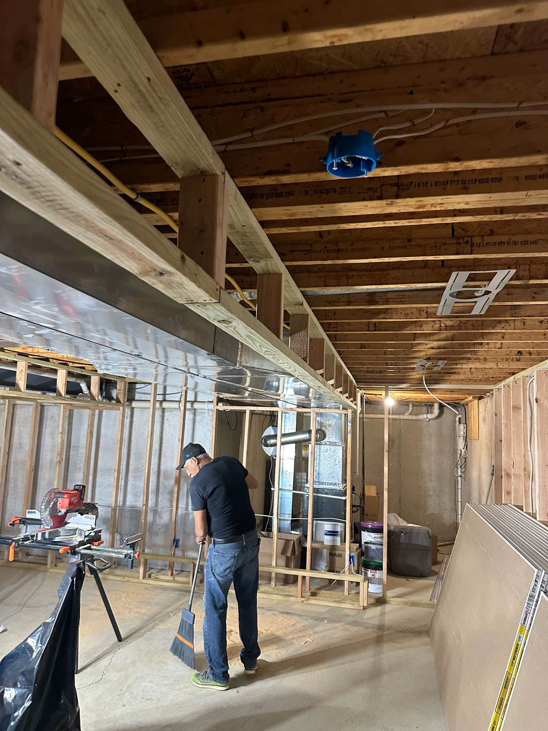 A person sweeping the floor in an unfinished basement with exposed beams, ducts, and construction materials.