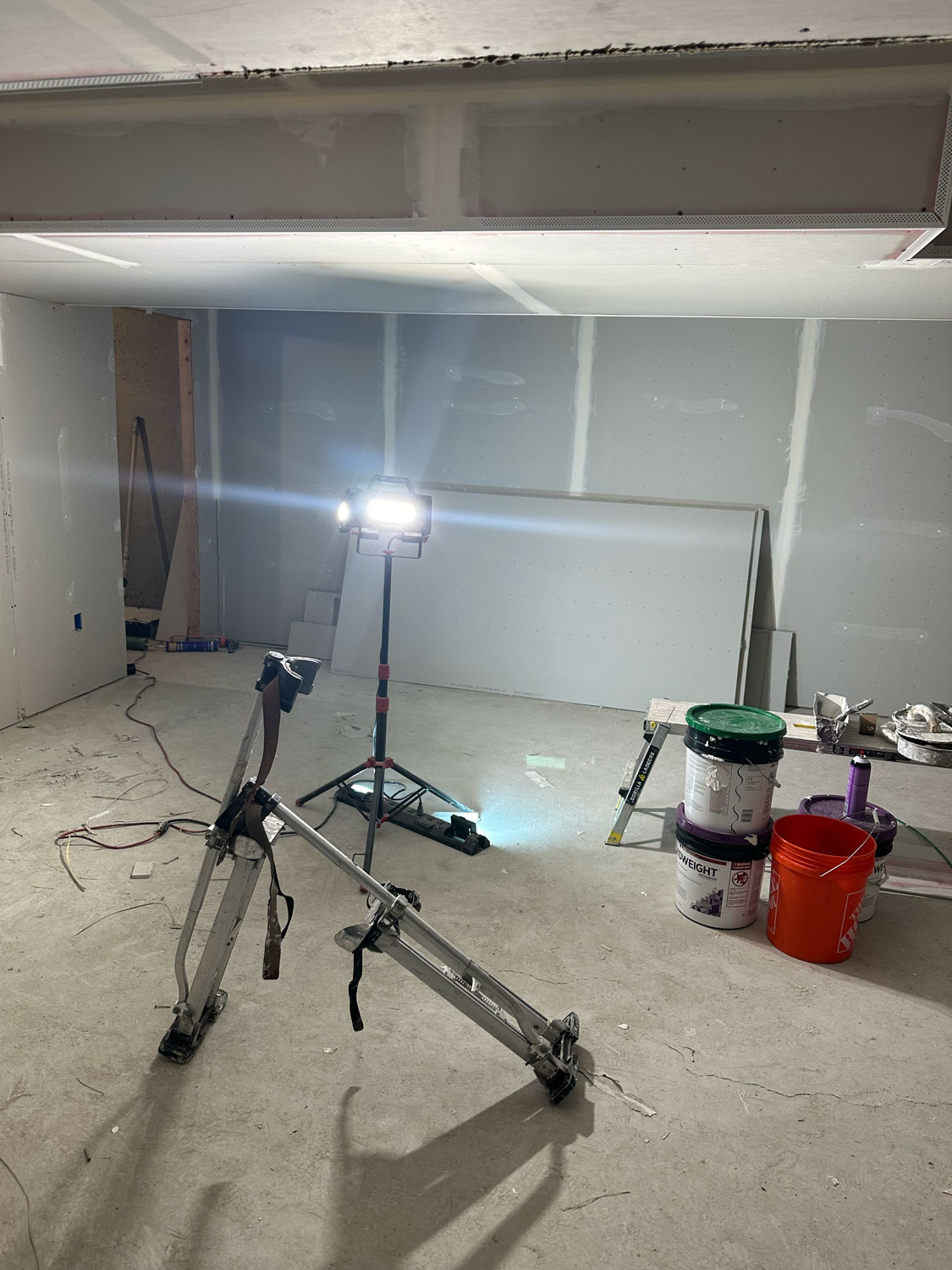 A construction site with drywall, work lights, stilts, paint cans, buckets, and tools on a concrete floor.