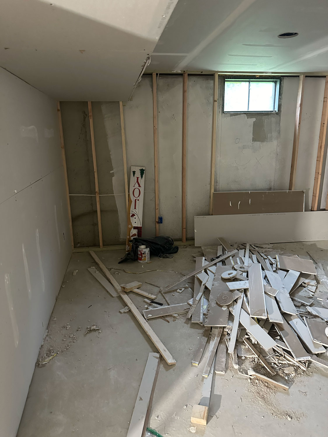 Room under construction with exposed wall studs, drywall sheets, debris, and a small window letting in light.