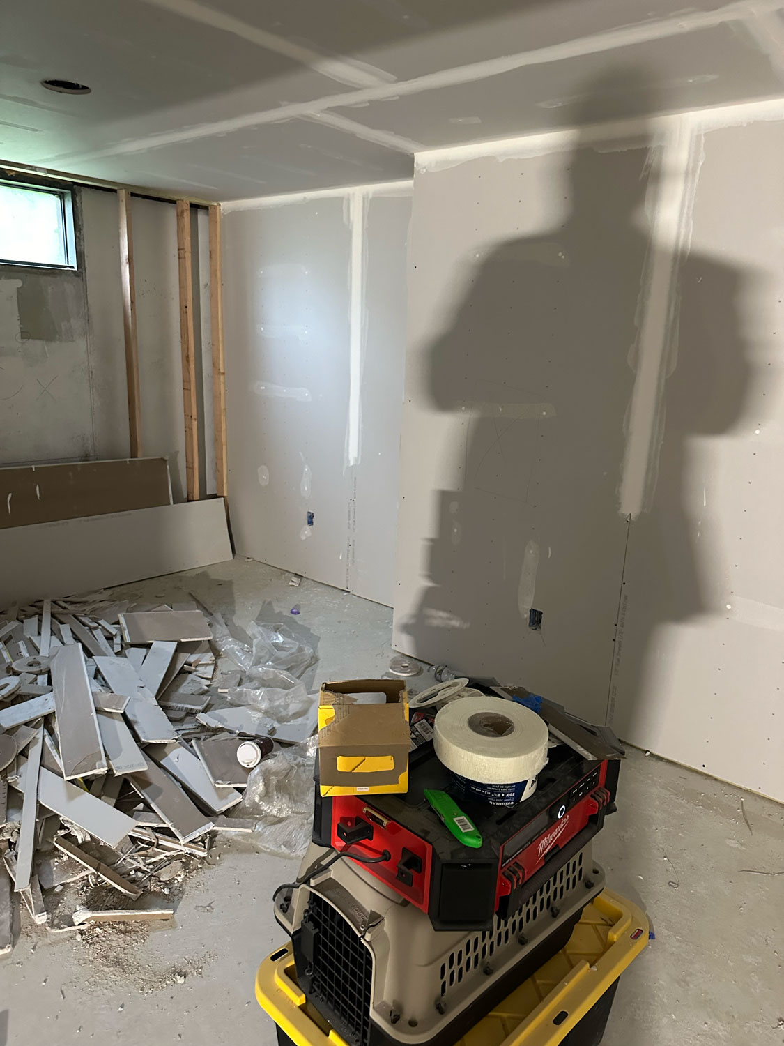 Room under construction with drywall, debris on floor, tools, and a large shadow cast on the unfinished walls.