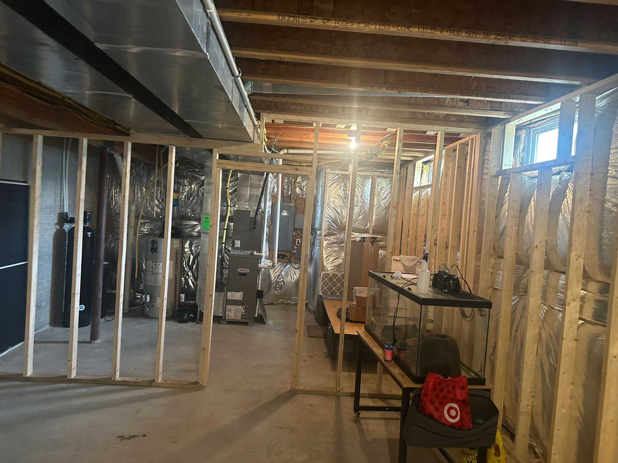 Unfinished basement with exposed framing, HVAC units, a table, and storage boxes under a small window.