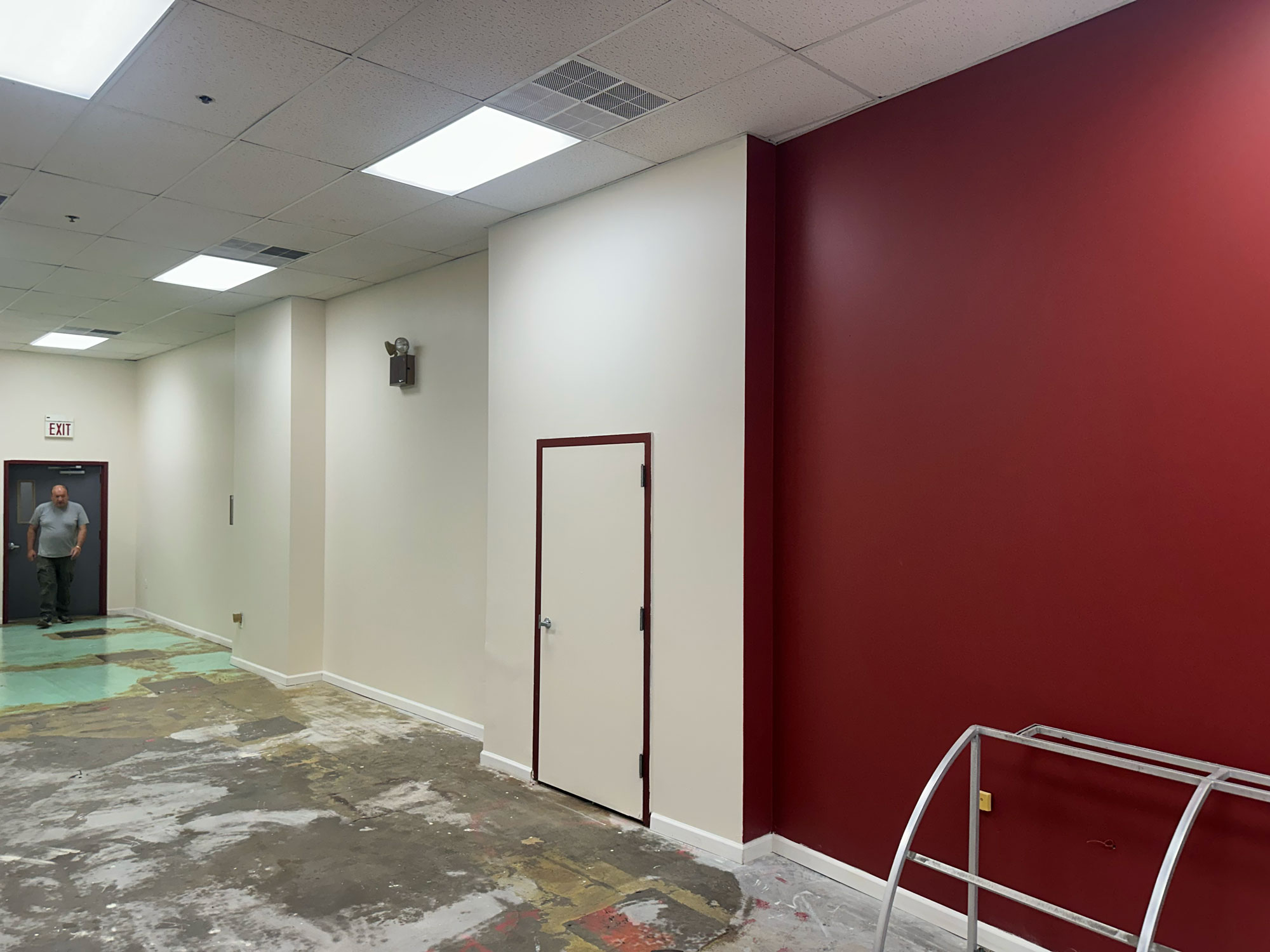 A man walks in a partially renovated room with a red accent wall, white door, and exposed flooring.
