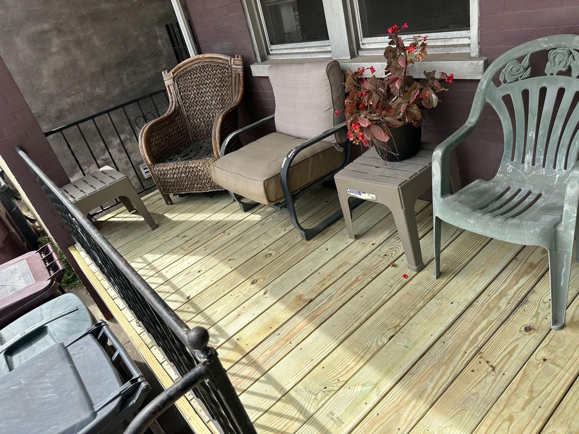 Three chairs and a small table with a potted plant on a wooden porch next to trash bins and a metal railing.