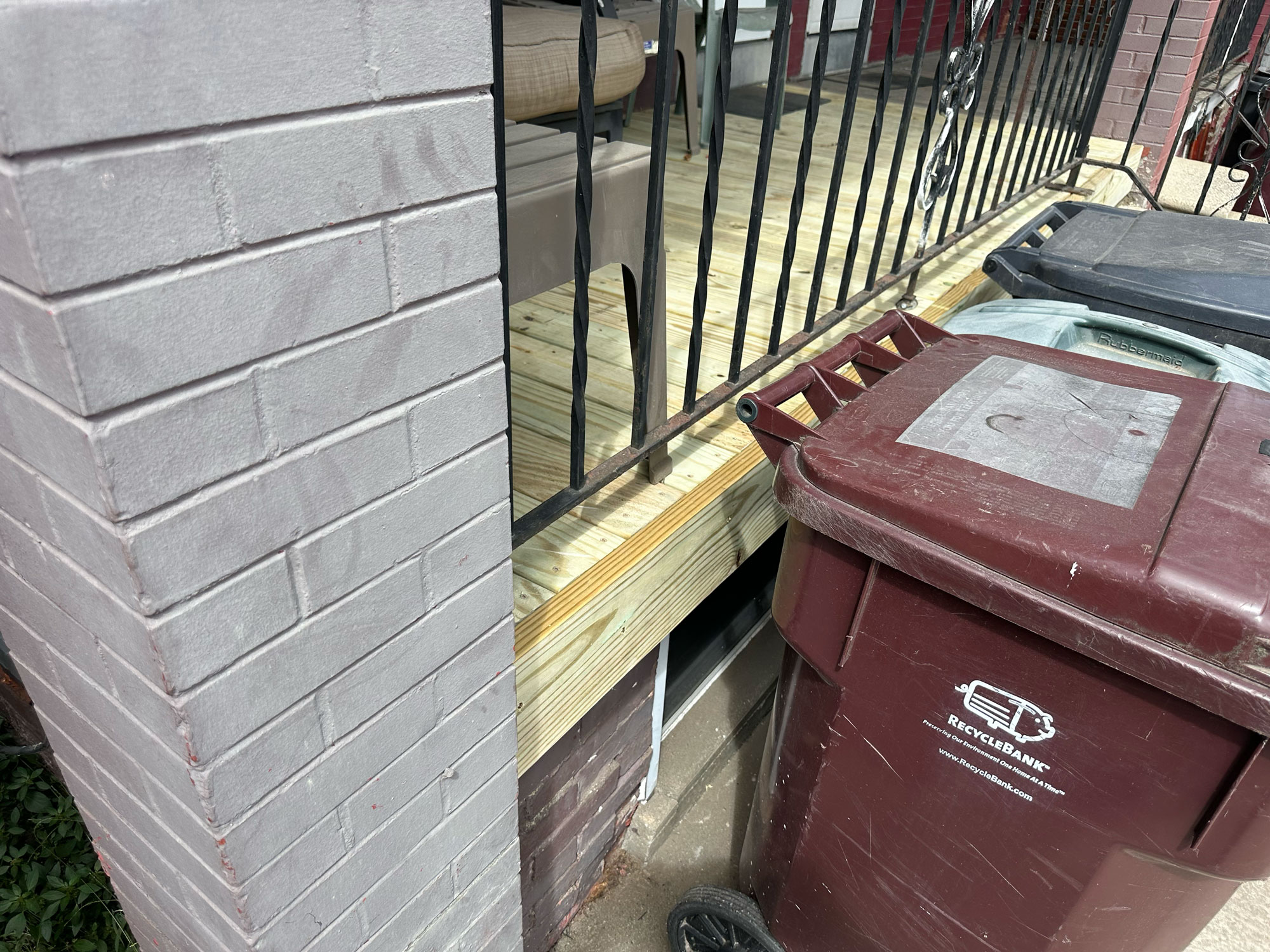 Close-up of a new wooden porch deck beside a brick wall and black railing, with recycling bins in front.