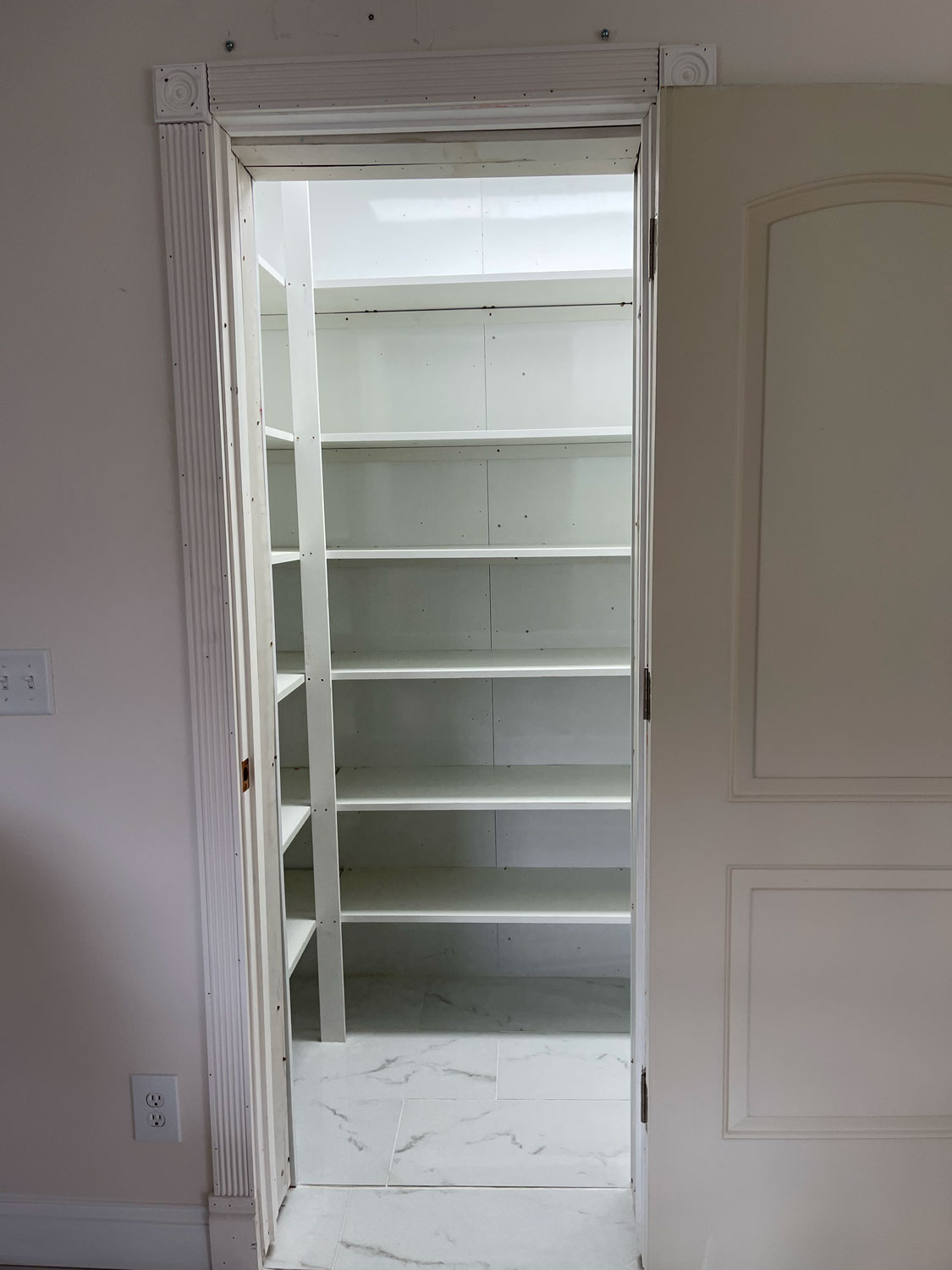 Open door revealing an empty white pantry with shelves and a tile floor.