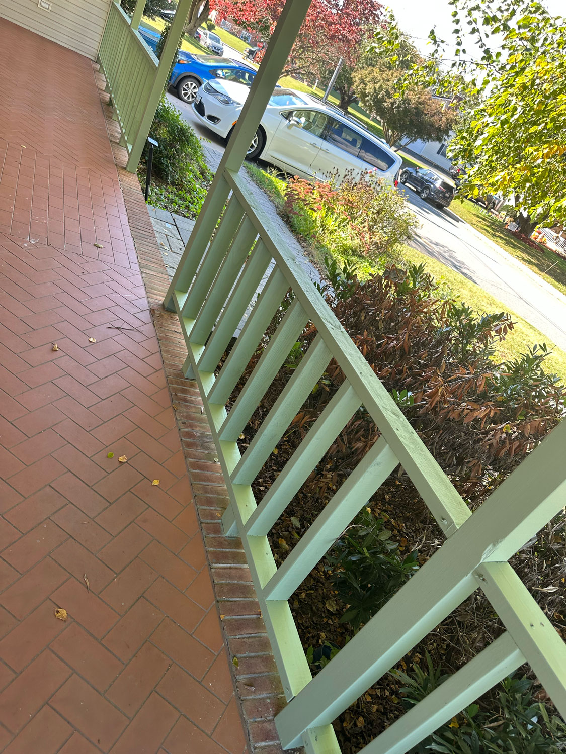 View from a porch with green railing, brick floor, and cars parked along a leafy suburban street.