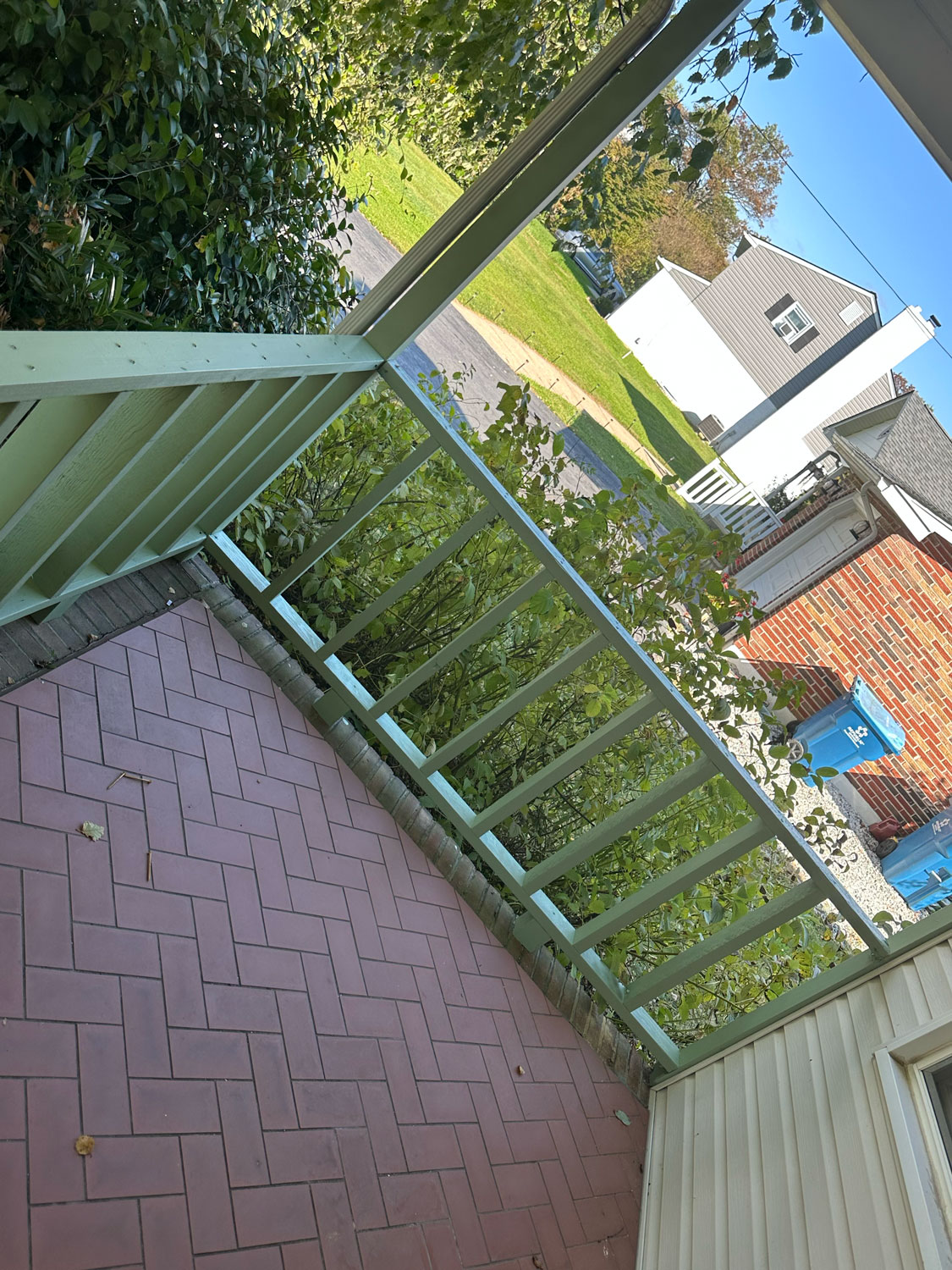View from a porch with green railing, brick-patterned floor, bushes, and houses across the street.