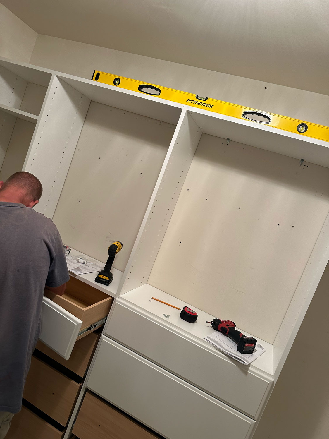A person installs drawers in a white closet unit with tools and a level placed on top.