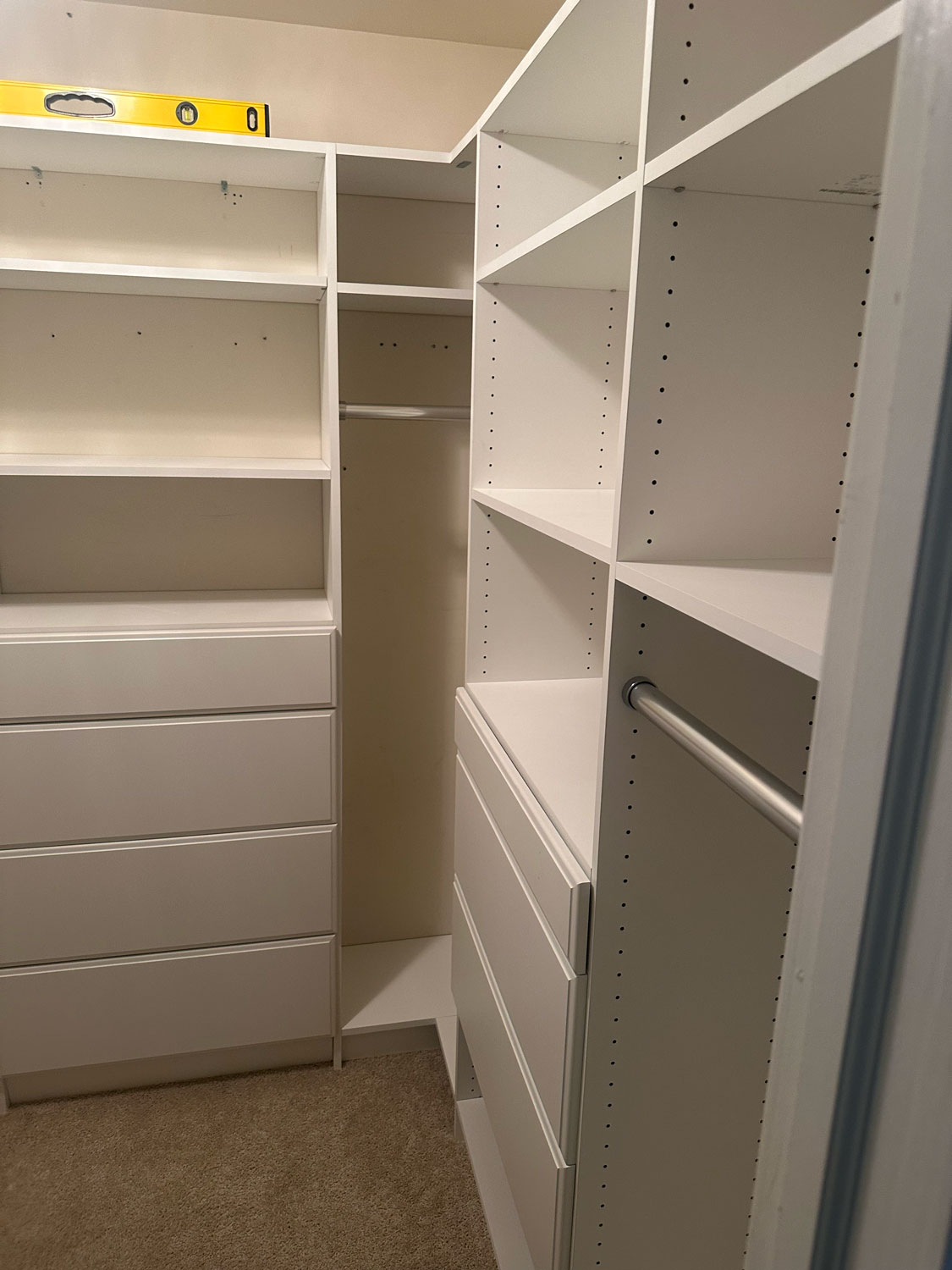 White walk-in closet with shelves, drawers, and hanging rods, with a yellow level on the top shelf.