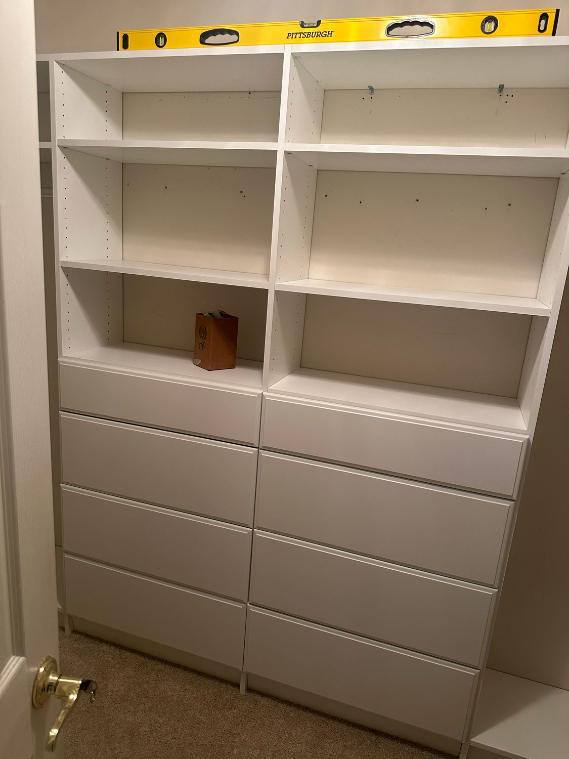 White shelving units, one tilted, with a yellow level tool on top and a small box on a shelf.