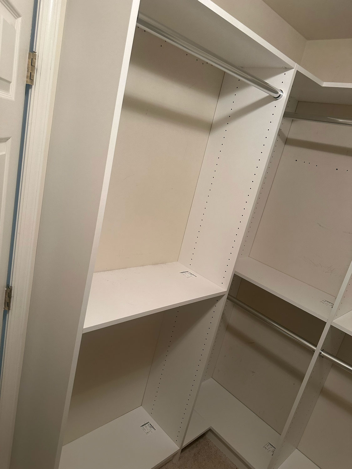 White built-in closet shelves and rods in an empty walk-in closet with beige carpeted floor.
