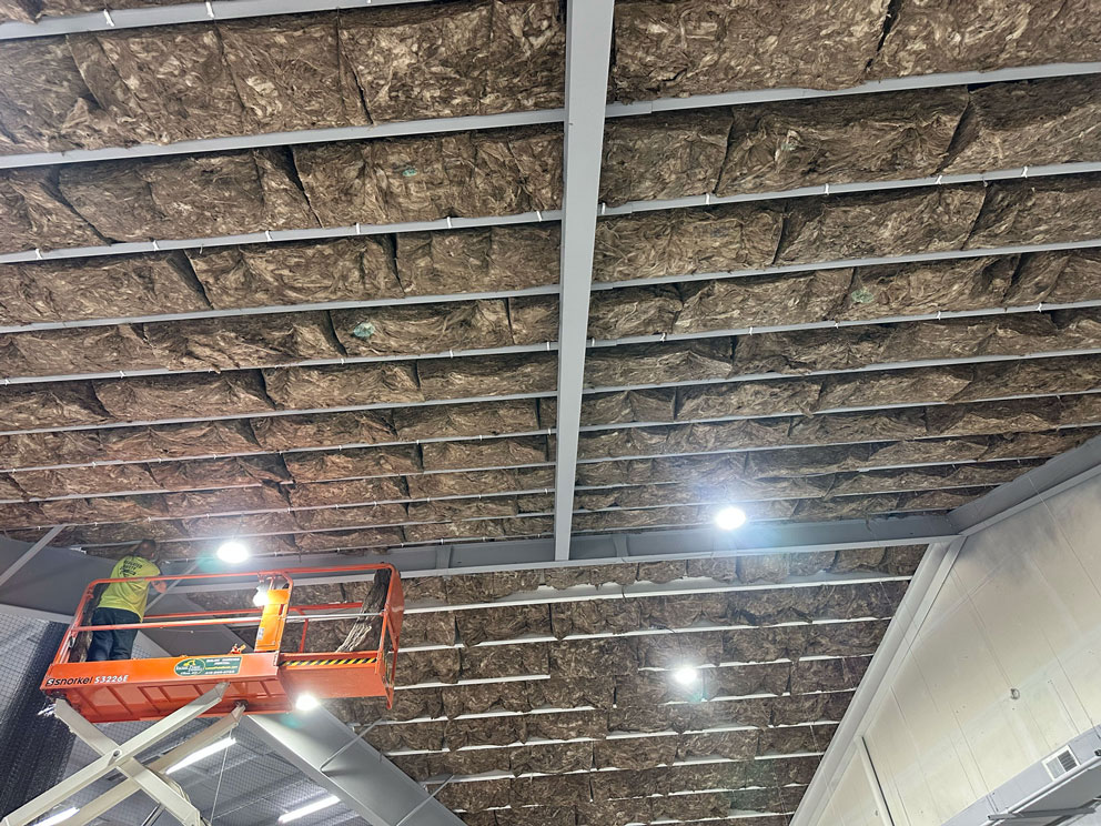 A worker on a lift installs insulation panels in the ceiling of a large industrial building.