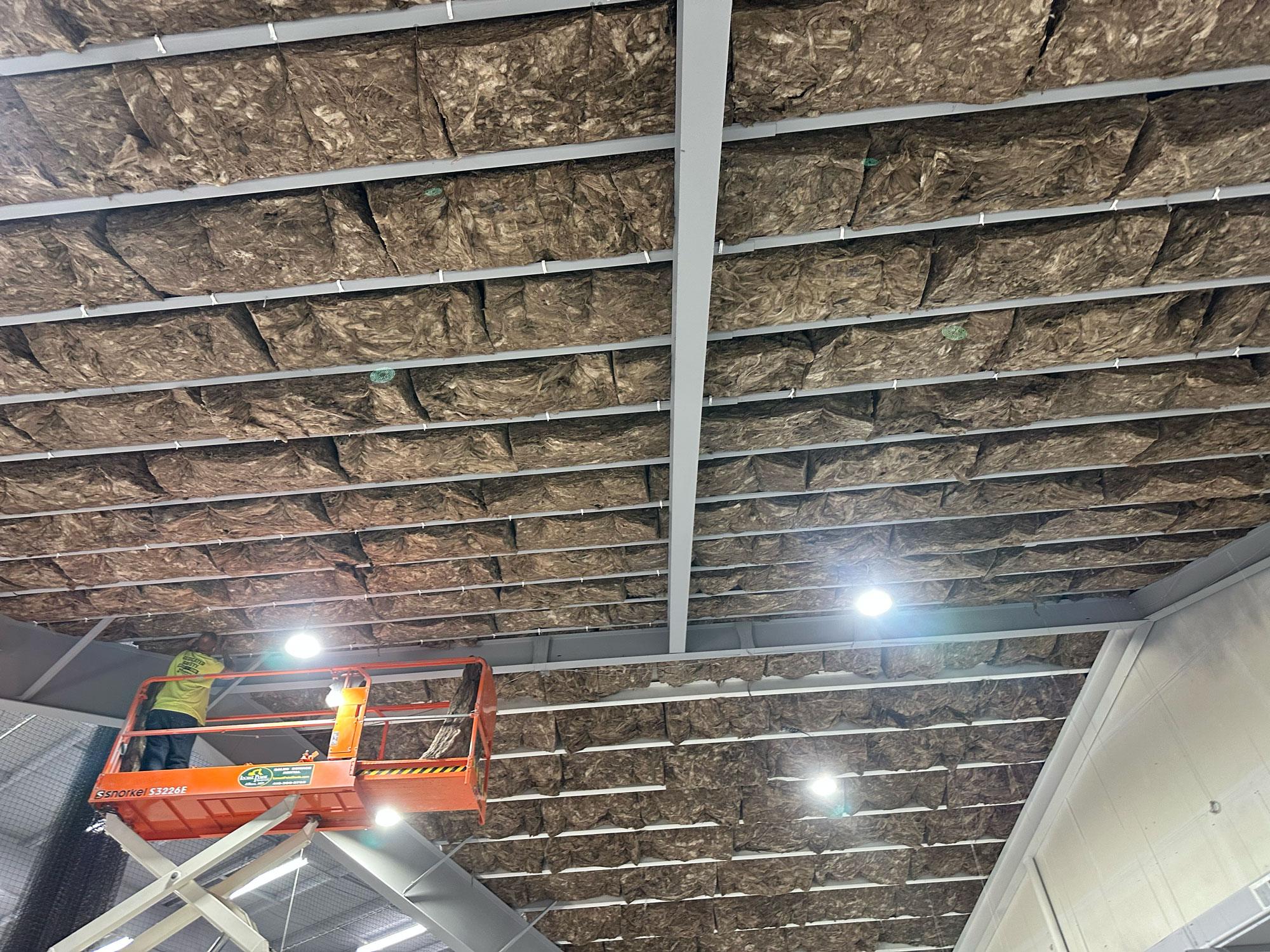 Worker on lift installing insulation panels on a large warehouse ceiling with exposed framework and lights.