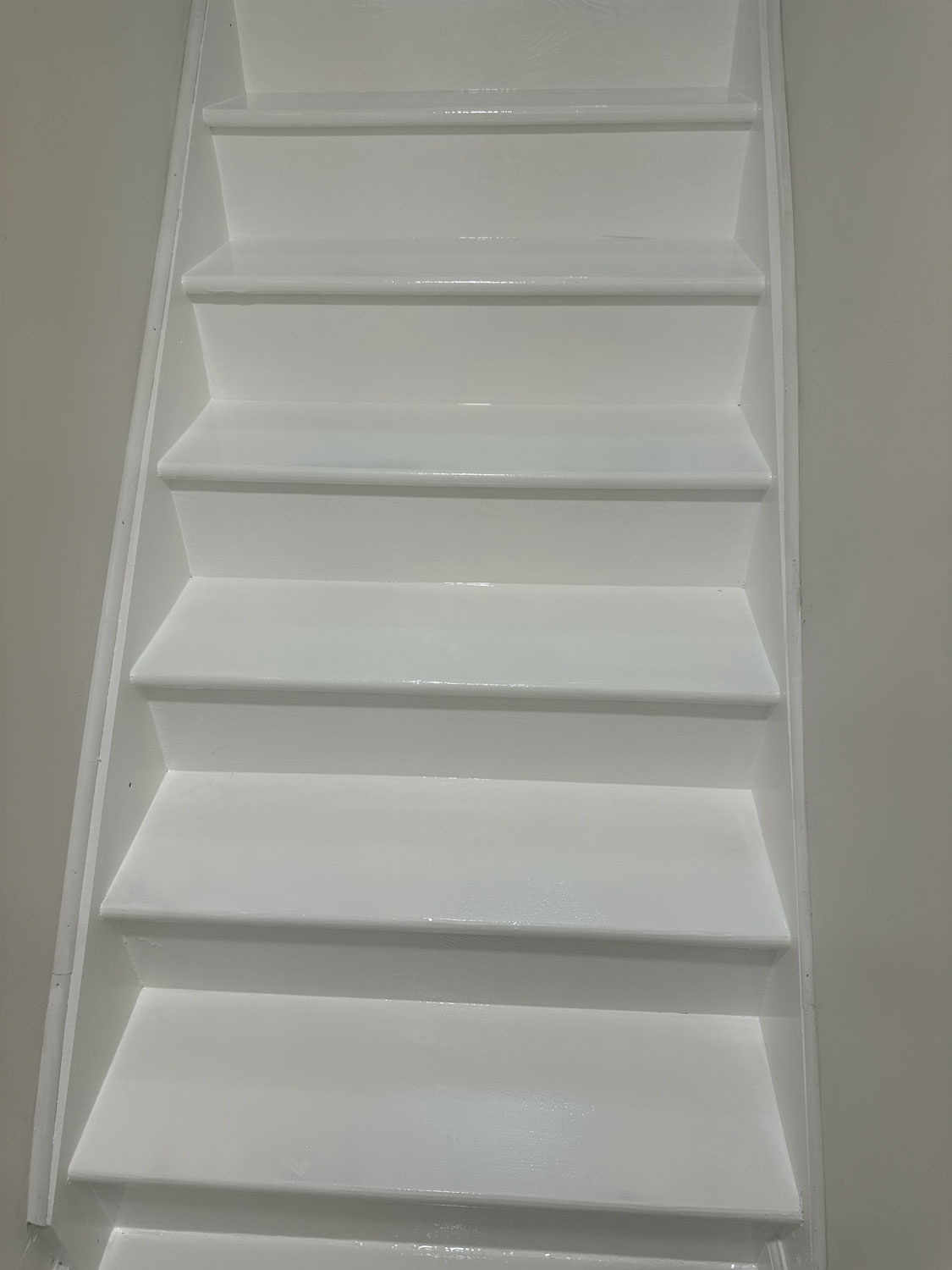 White, steep staircase with shiny steps against a light grey wall, viewed from the bottom looking up.