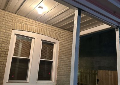 Lit porch at night with a double window, brick wall, wooden fence, and metal railing.