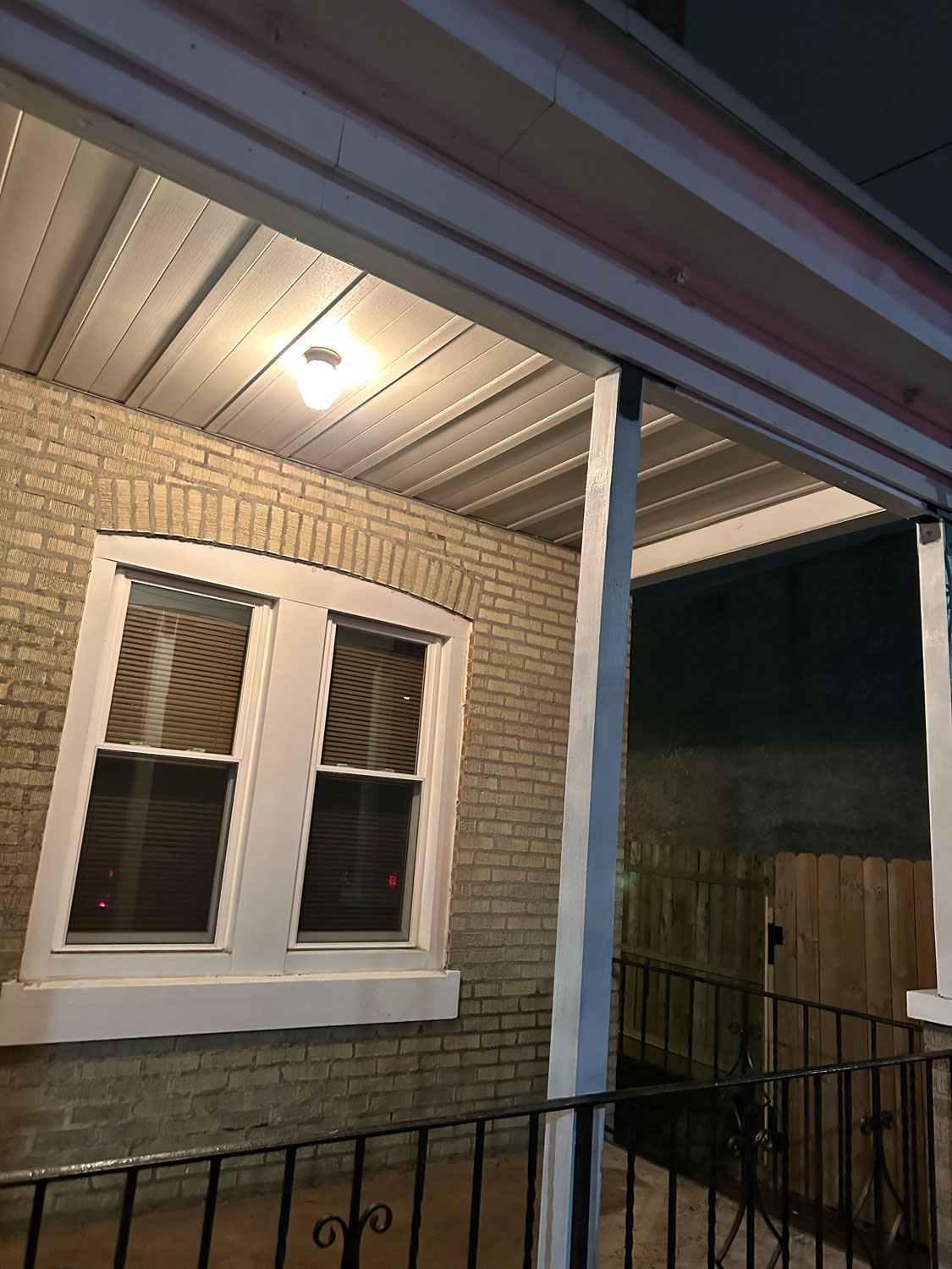 Lit porch at night with a double window, brick wall, wooden fence, and metal railing.