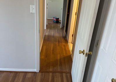 Open white door leading to a hallway with wooden floors and a bag visible in the distant room.