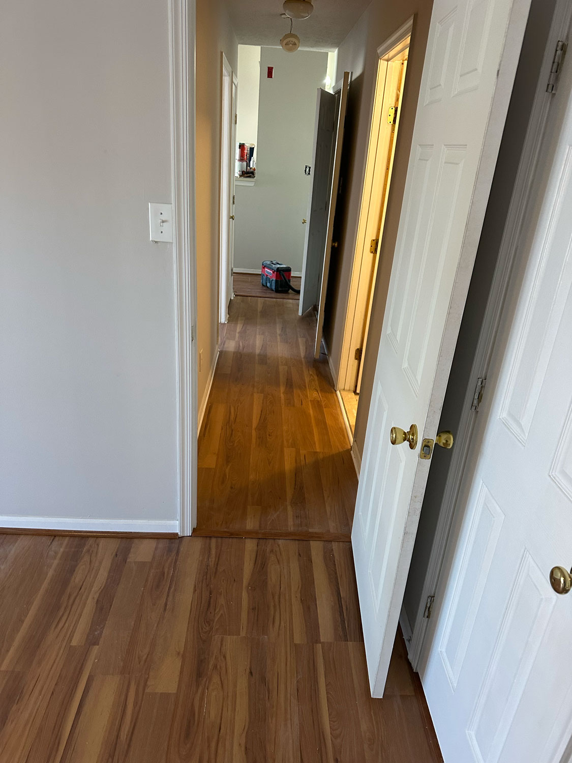 Open white door leading to a hallway with wooden floors and a bag visible in the distant room.