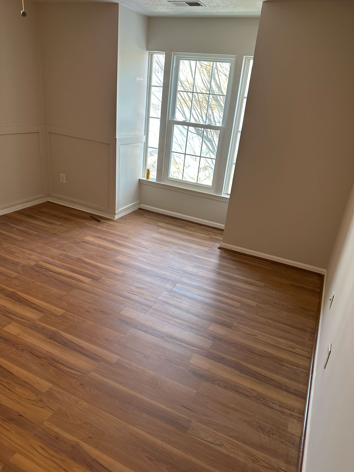 Empty room with wood flooring, large windows, and beige walls, sunlight streaming in through the glass.