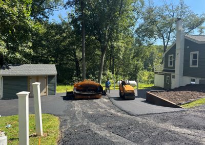 Two workers pave a driveway next to a gray house and garage on a sunny day with trees in the background.