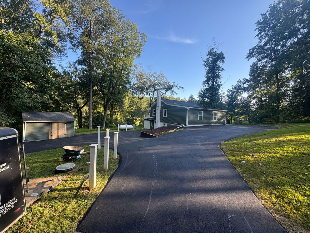 A paved driveway leads to a gray house surrounded by trees and green grass under a clear sky.