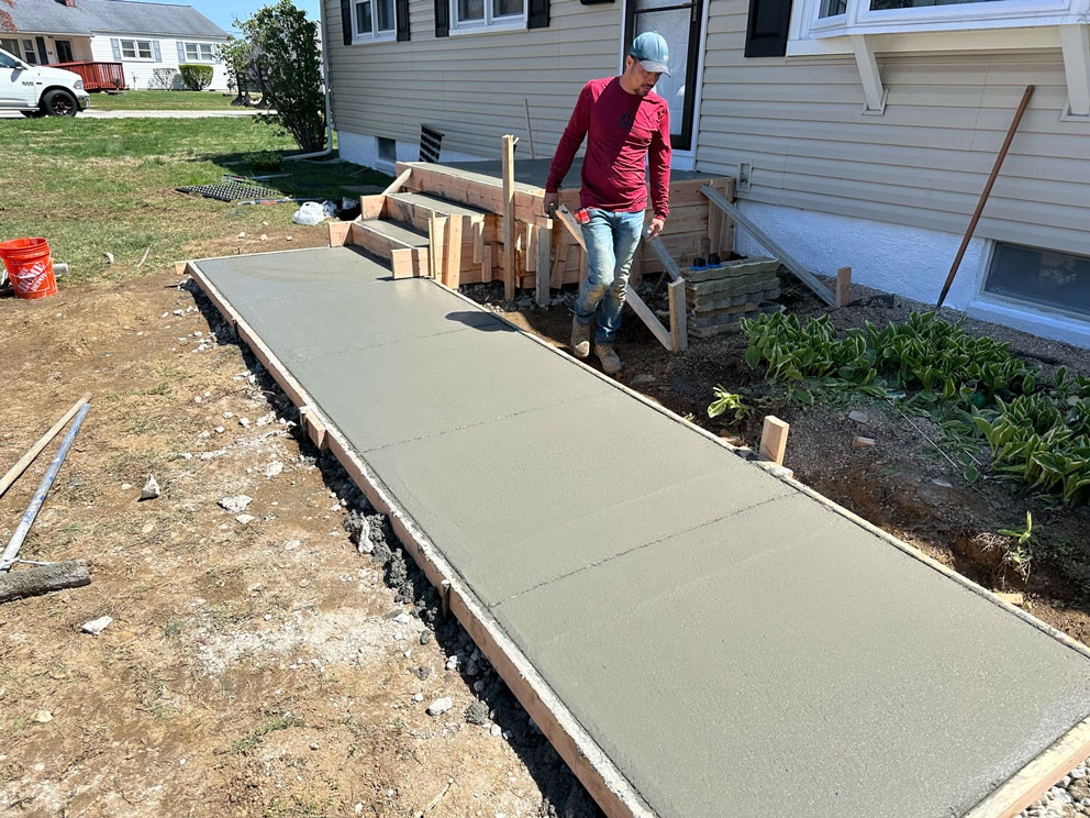 A person stands next to freshly poured concrete forming a sidewalk in front of a house.