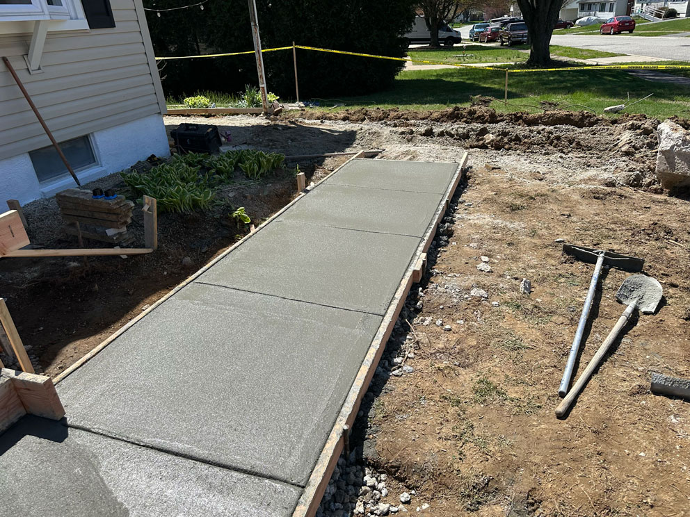 Freshly poured concrete sidewalk with tools and dirt around, next to a house and a grassy yard.
